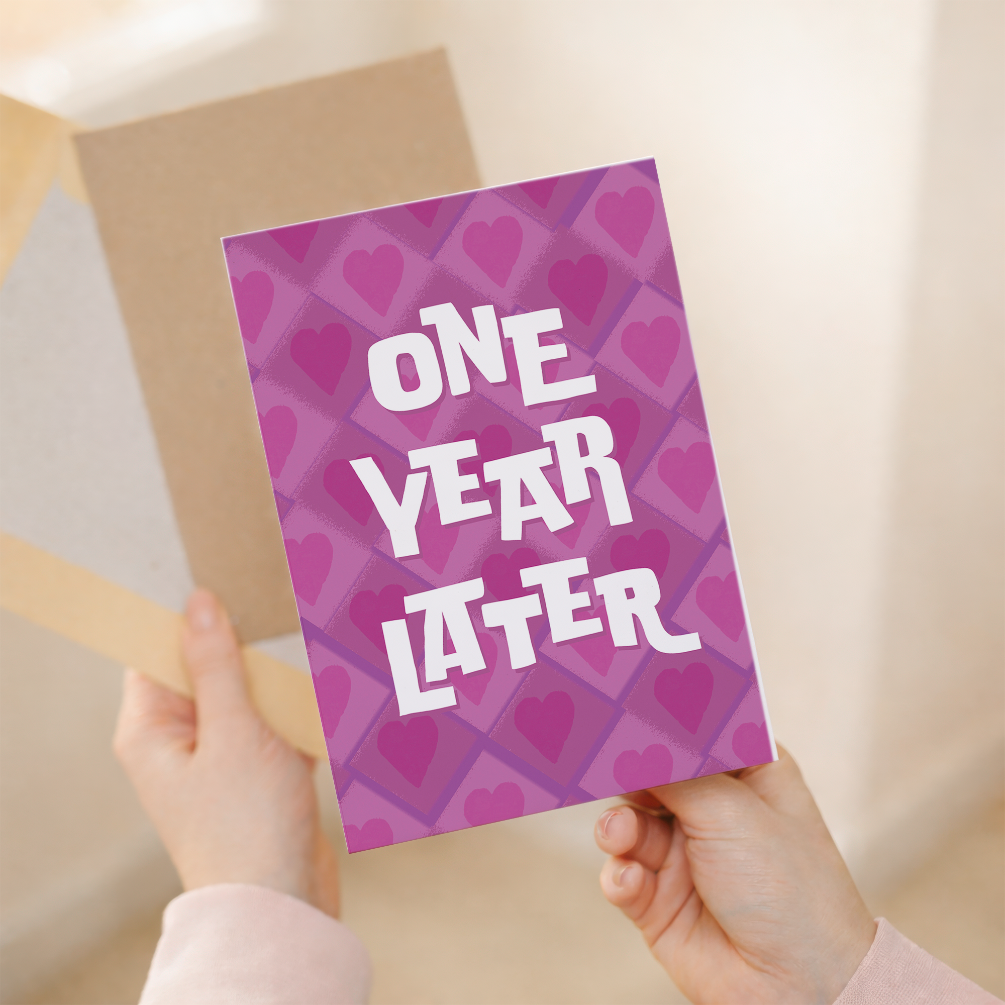 A simple and modern 5x7 inch 1st wedding anniversary greeting card featuring a clean design in pink, white, and purple on sustainable recycled cardstock, accompanied by a brown Kraft envelope.