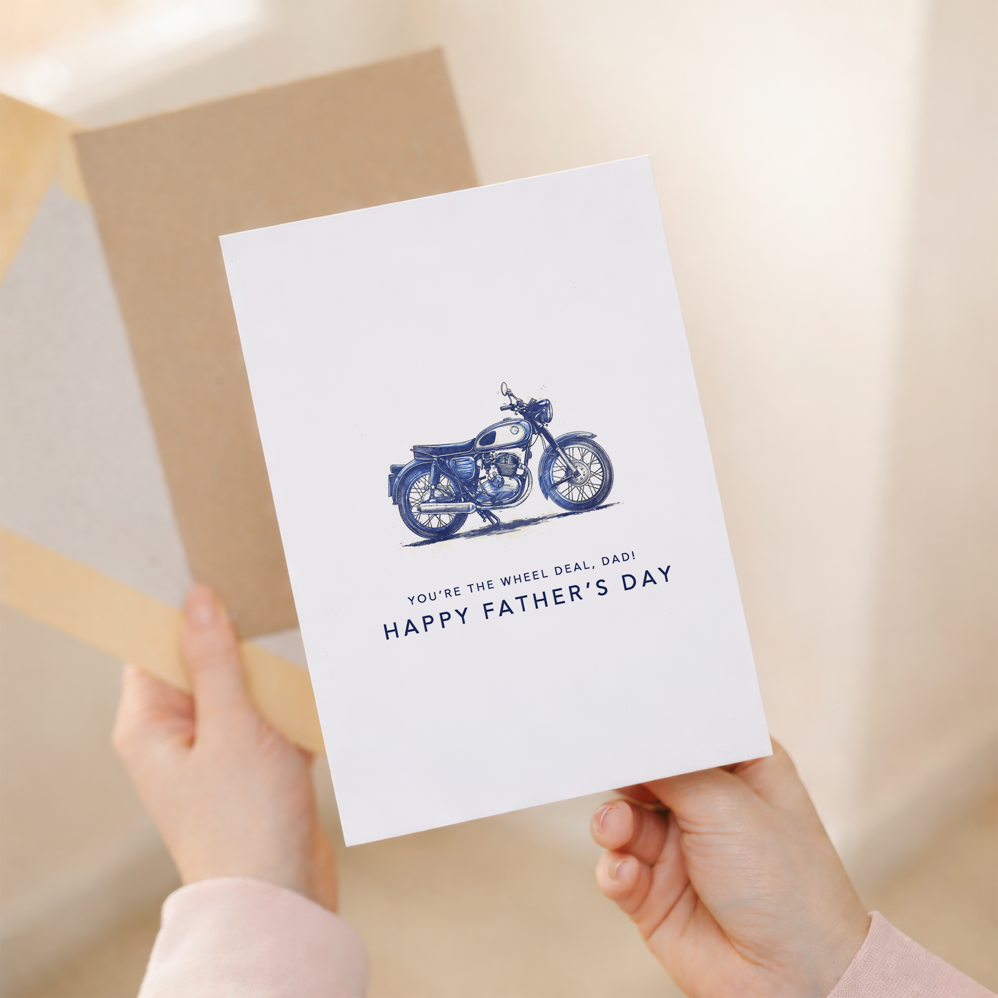 A simple and modern Father's Day greeting card for motorcycle enthusiasts featuring a minimalist blue motorbike illustration and the punny text You're the Wheel Deal Dad on a white background.
