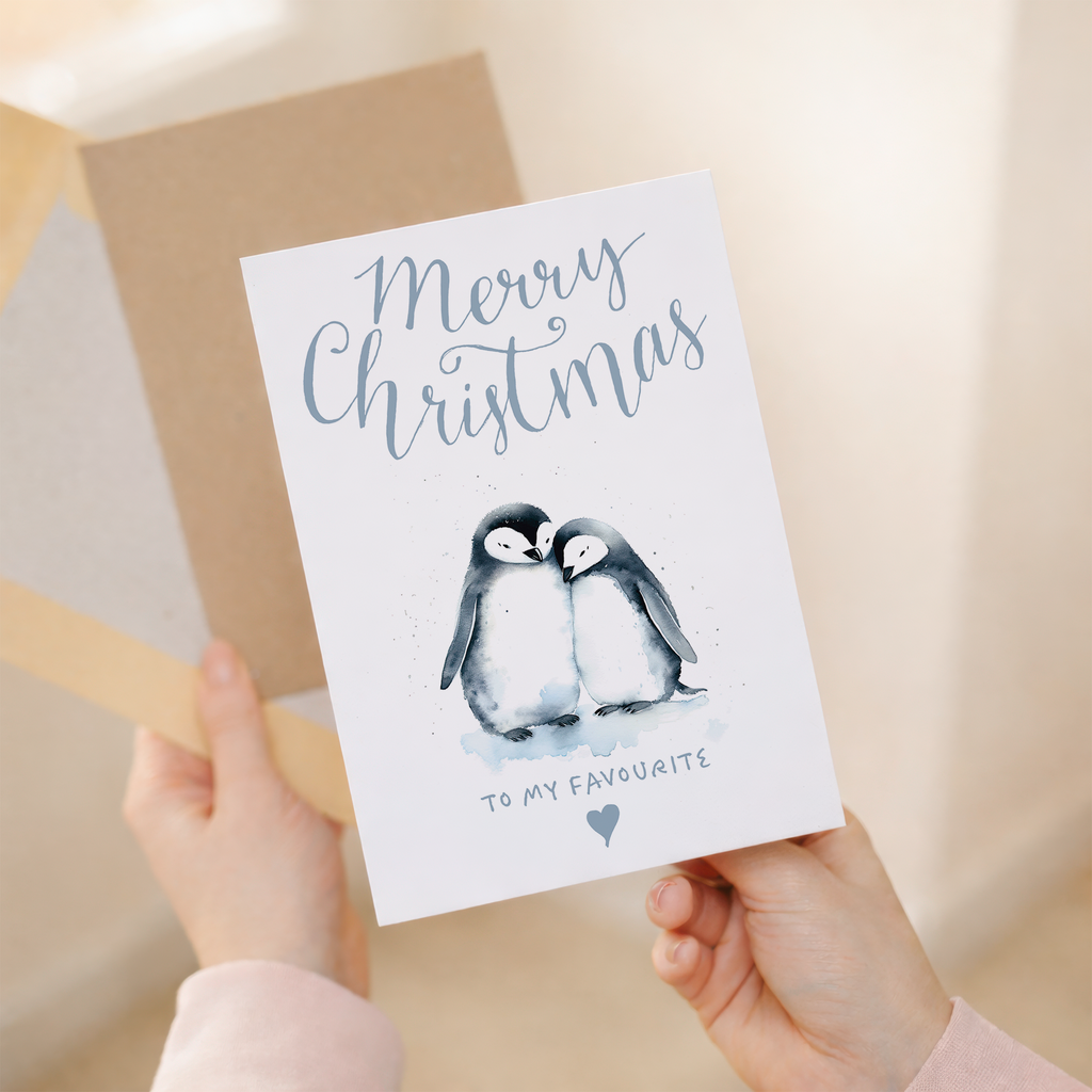 Illustration of two cute penguins wearing red scarves and Santa hats, standing on snow against a light blue background. One penguin holds a small gift. Text on the card reads: To the one I love at Christmas. The card is displayed next to a brown kraft paper envelope.