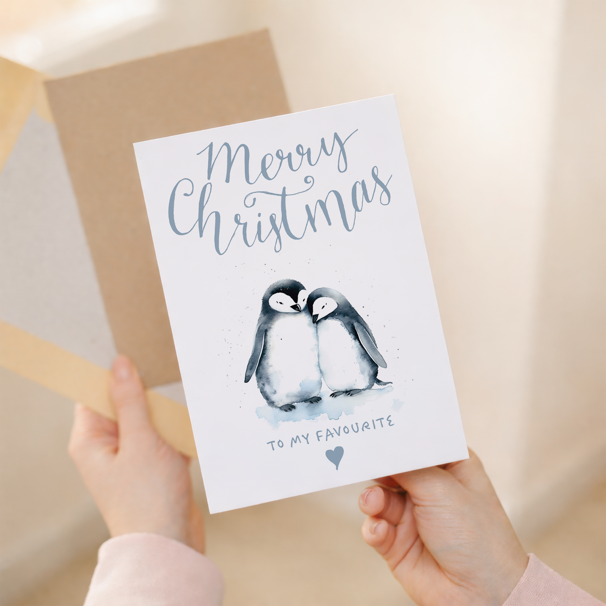 Illustration of two cute penguins wearing red scarves and Santa hats, standing on snow against a light blue background. One penguin holds a small gift. Text on the card reads: To the one I love at Christmas. The card is displayed next to a brown kraft paper envelope.