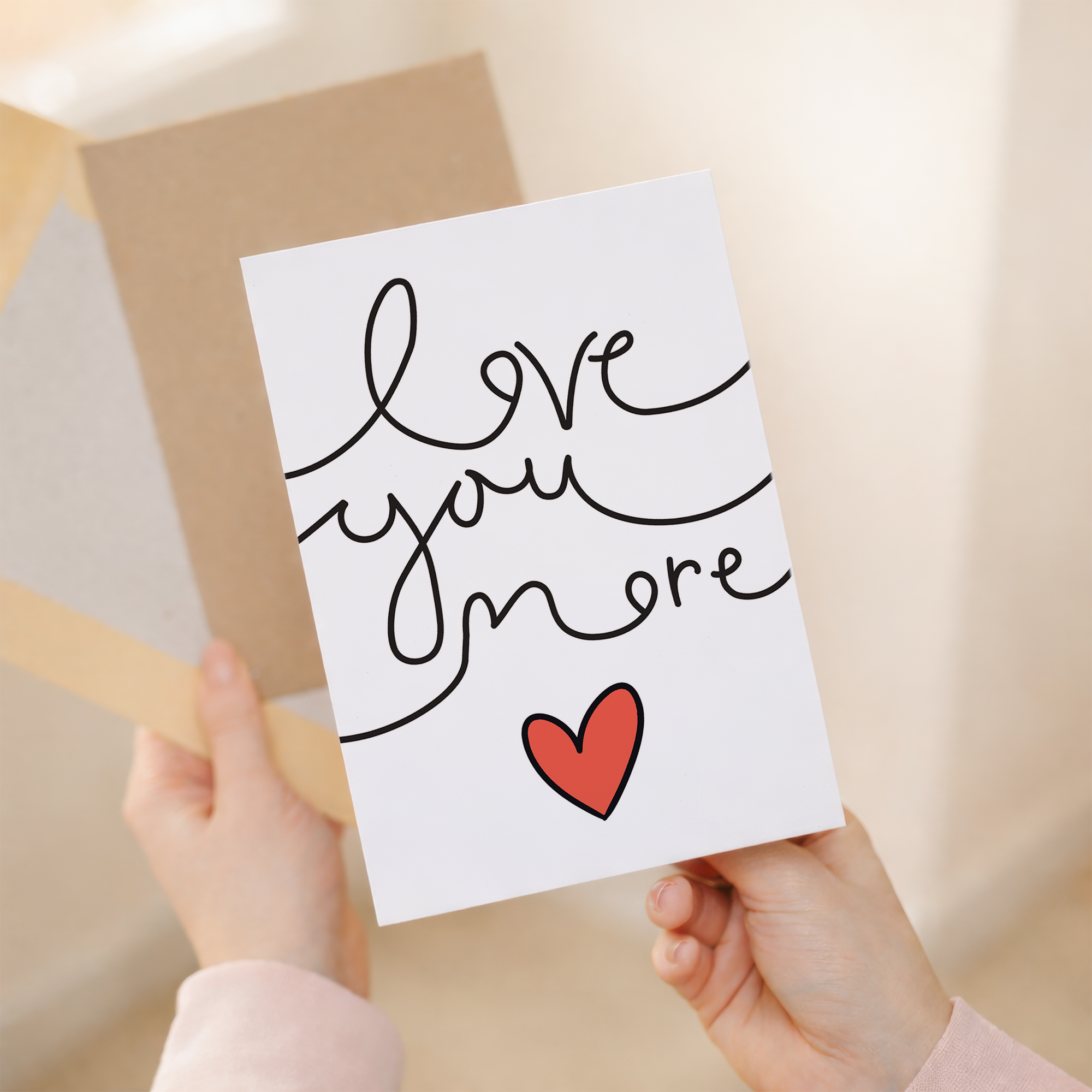 Minimalist Valentine's Day greeting card featuring the text Love You More in a stylish script on sustainable recycled cardstock, accompanied by a brown Kraft envelope.