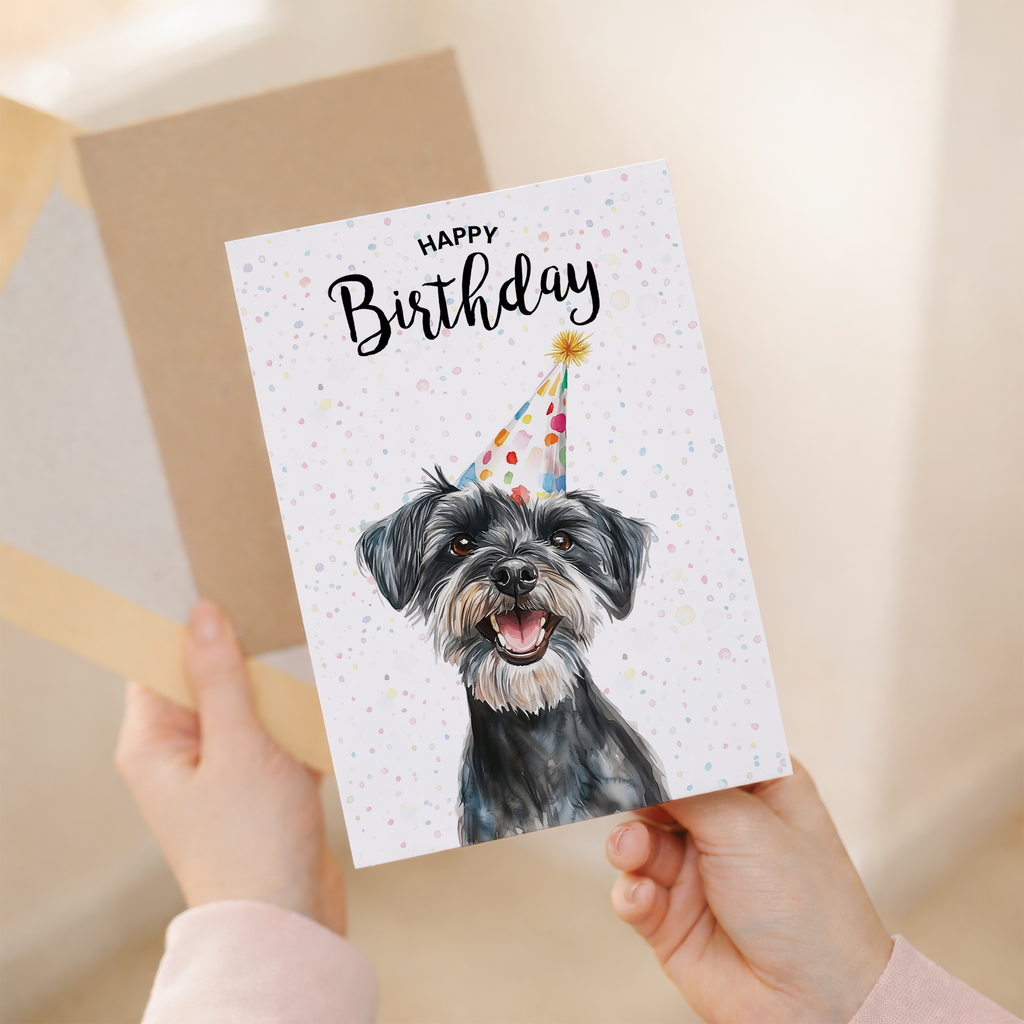 A watercolour illustration of a Schnauzer dog with a celebratory party hat on a white birthday greeting card.