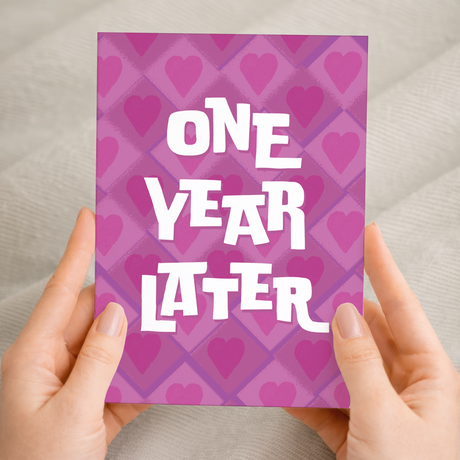 A simple and modern 5x7 inch 1st wedding anniversary greeting card featuring a clean design in pink, white, and purple on sustainable recycled cardstock, accompanied by a brown Kraft envelope.