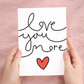 Minimalist Valentine's Day greeting card featuring the text Love You More in a stylish script on sustainable recycled cardstock, accompanied by a brown Kraft envelope.