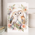 Illustrated anniversary greeting card featuring two cute owls sitting on a branch surrounded by soft green floral and leaf patterns on a beige background.