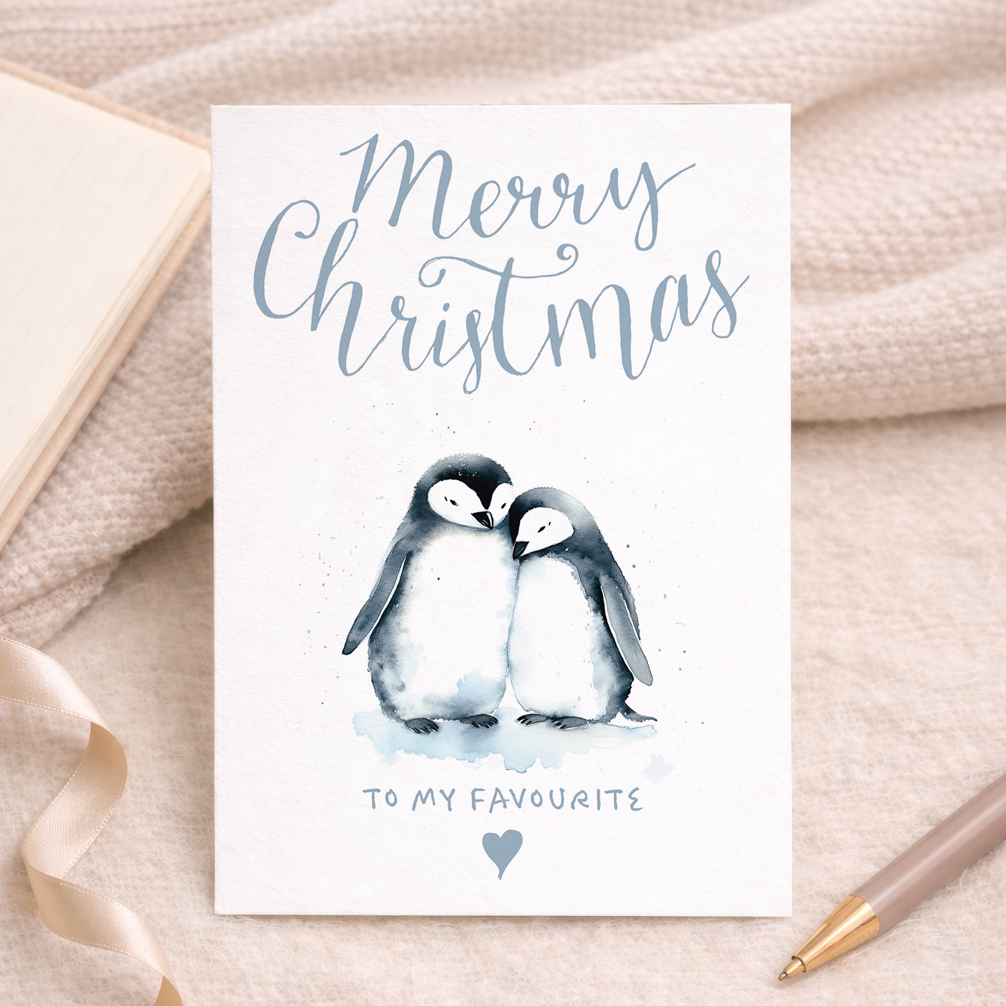 Illustration of two cute penguins wearing red scarves and Santa hats, standing on snow against a light blue background. One penguin holds a small gift. Text on the card reads: To the one I love at Christmas. The card is displayed next to a brown kraft paper envelope.