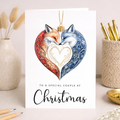 A romantic Christmas card featuring an illustration of two foxes and a red love heart inside a festive bauble design.