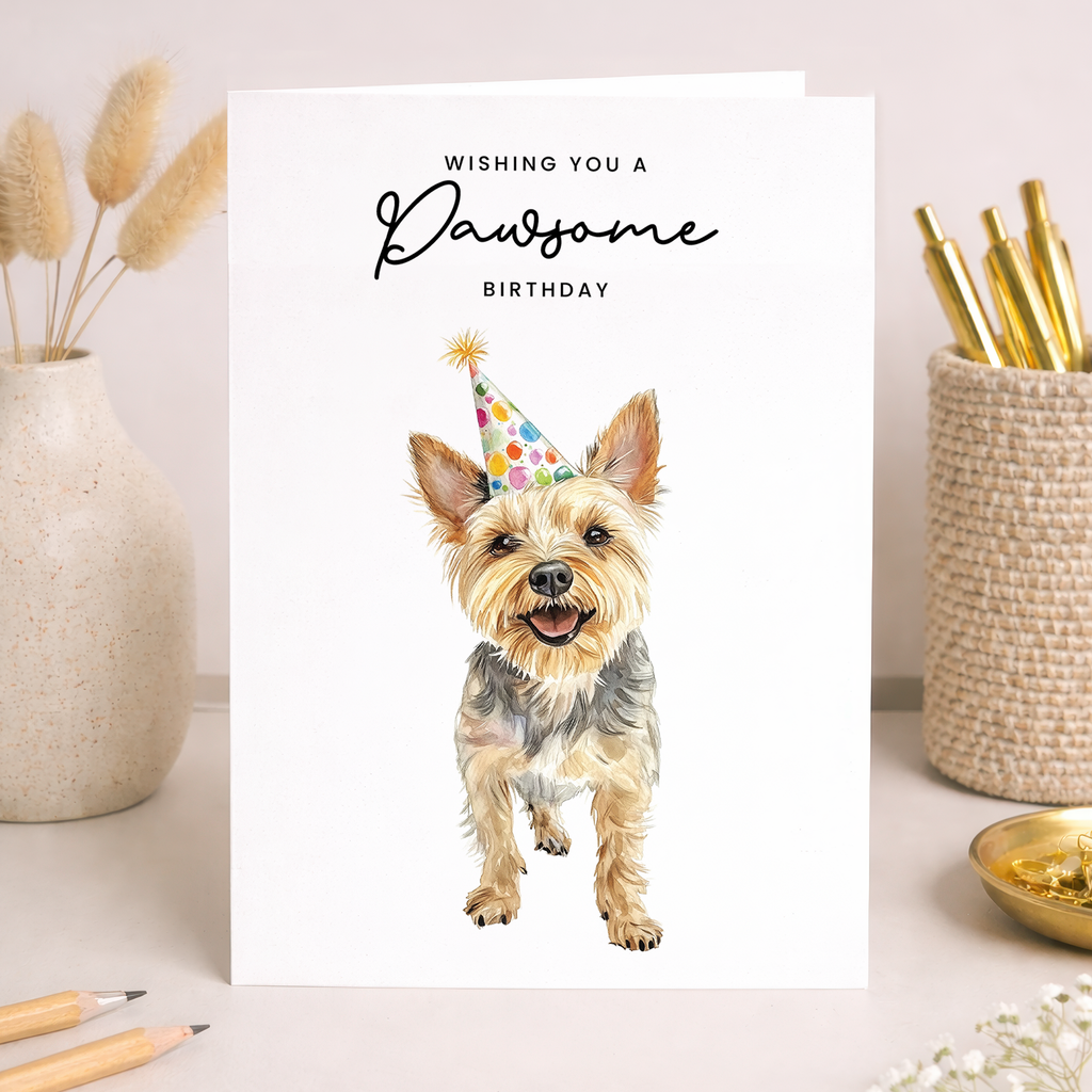A cute Yorkshire Terrier wearing a colorful party hat on a white background with the text Wishing You a Pawsome Birthday.