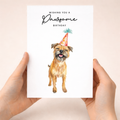 A cute Border Terrier wearing a colorful party hat on a minimalist birthday greeting card with the text Wishing You a Pawsome Birthday.