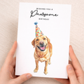 A happy yellow labrador retriever wearing a colorful, striped party hat, featured on a 5x7 inch birthday greeting card with the caption Wishing You a Pawsome Birthday.